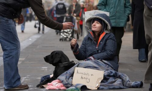 a young woman on the street sitting in a pile of blankets with her dog and a sign that says "homeless and hungry;" a person off screen offers her a cup of coffee